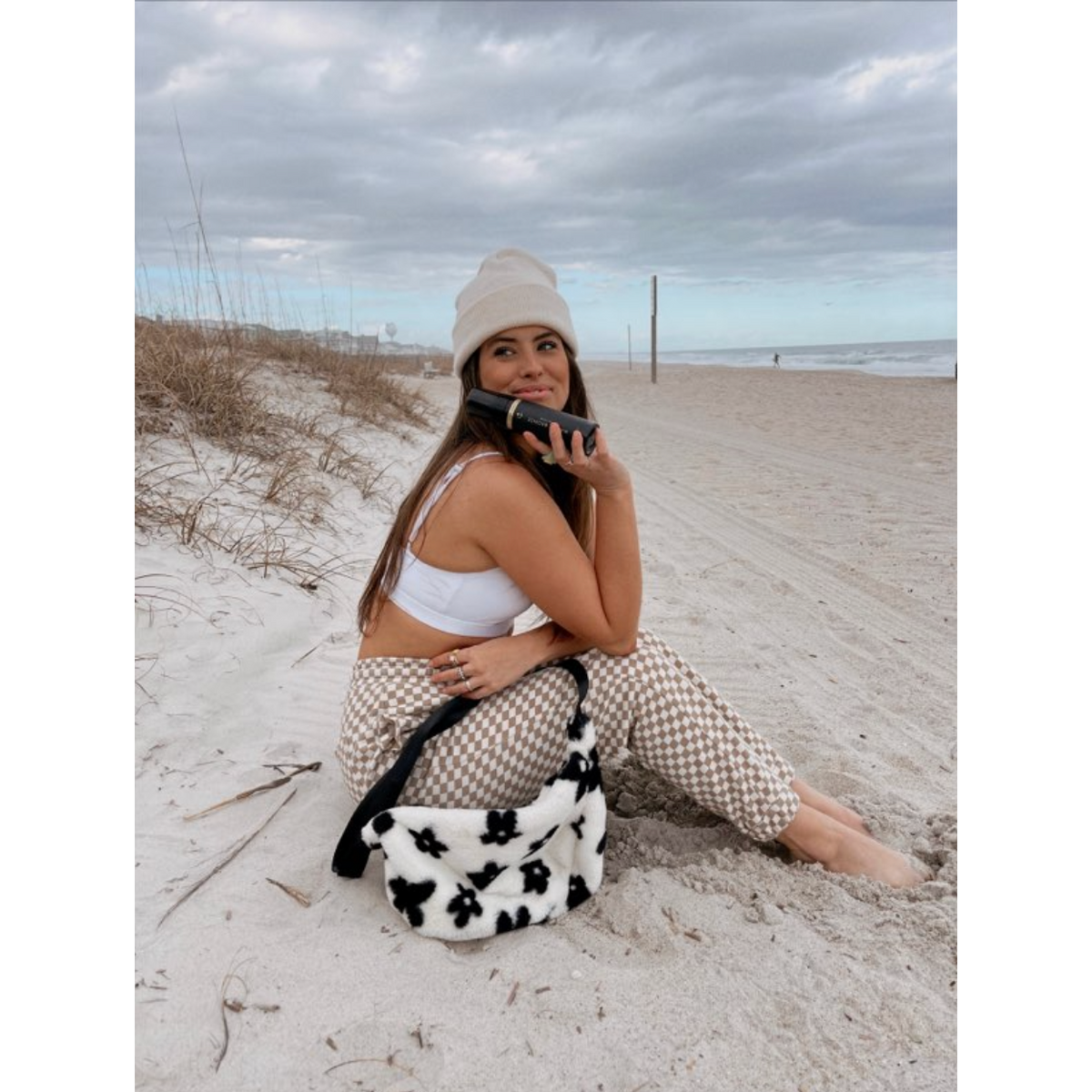 Influencer posing with Malibu Bronze Sunless Tan Bottle at the beach while wearing a hat by the ocean while on the sand. 